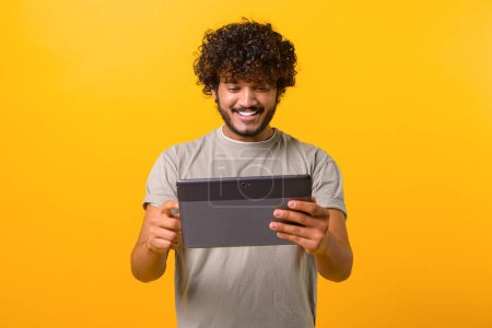 Foto de Positive handsome Indian young curly man using digital tablet for web surfing or remote working standing isolated on yellow background, foreign male student using computer app for studying online - Imagen libre de derechos