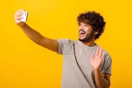 Téléchargez les photos : Waist up view of the young Indian man waving with hand while chatting via cell phone or making selfie isolated on yellow background - en image libre de droit