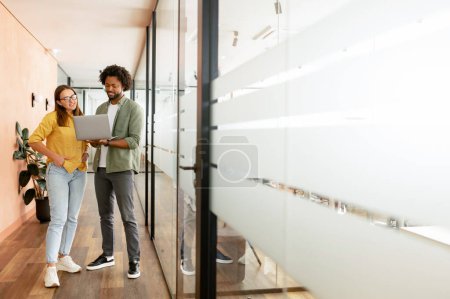 Foto de Full-length photo of man and woman looking at the laptop screen while standing in the glass office walkhall, coworkers discussing about project, two colleagues brainstorming together - Imagen libre de derechos