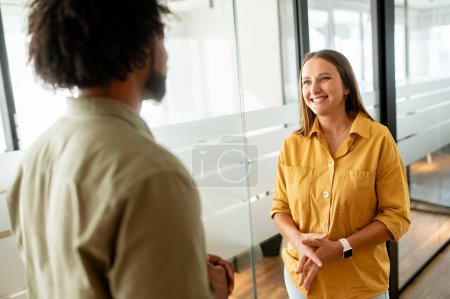 Foto de Dos colegas tienen una agradable conversación de pie en el pasillo de la oficina moderna de espacio abierto, sonriendo y disfrutando de un ambiente de trabajo amigable, mujer sonriente escuchando a su compañero de trabajo masculino - Imagen libre de derechos