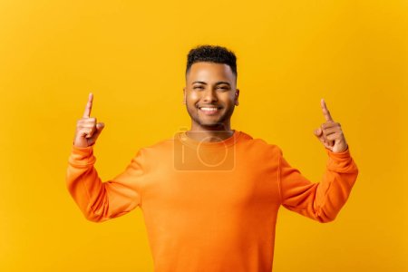 Foto de Attractive indian man with overjoyed facial expression pointing up with both fingers, presenting copy space. Indoor studio shot isolated on yellow background - Imagen libre de derechos