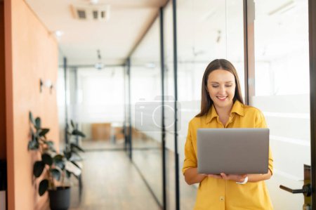 Photo for Smart and intelligent businesswoman wearing smart casual yellow shirt using laptop indoors, female office employee holding computer and typing message, smiling, enjoying her job - Royalty Free Image