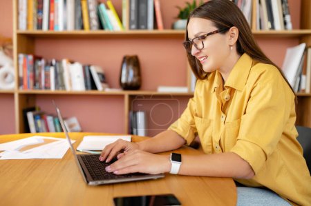 Photo for Cheerful female employee works with laptop in the modern office. Young positive woman in informal wear typing on the keyboard, a journalist is writing an article, female student in the library - Royalty Free Image