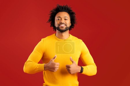 Photo for Handsome smiling African American curly young man showing thumbs up, ok gesture on red background, charismatic guy in yellow pullover expresses approval and support, look at the camera - Royalty Free Image