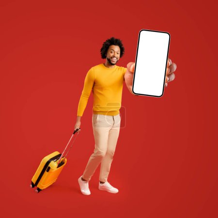 Photo for Happy smiling 30s curly guy holding yellow suitcase and showing smartphone with empty blank screen isolated on red background, presentation of mobile app for booking or renting, full length - Royalty Free Image