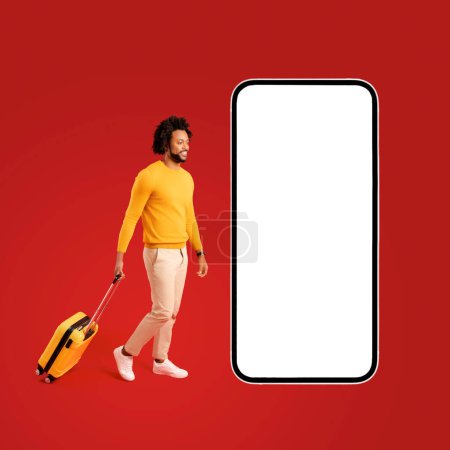 Téléchargez les photos : Mobile app for travelers. Curly African-American man with yellow luggage suitcase walking toward the giant huge smartphone with empty display, copy space, mockup - en image libre de droit