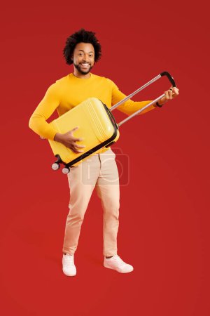Foto de Overjoyed 30s guy holding yellow suitcase and pretend playing guitar, ready for adventure and holiday, dancing and fulling around isolated on red background, travel and vacation concept. Full length - Imagen libre de derechos