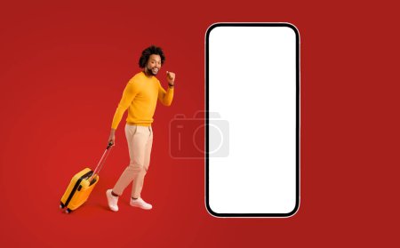 Téléchargez les photos : Le gars afro-américain excité portant une valise jaune court vers un énorme téléphone portable, un homme multiracial ravi se précipitant vers des vacances et des aventures. App pour les voyageurs - en image libre de droit
