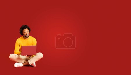 Foto de Hombre rizado afroamericano guapo confiado sentado en el suelo en pose de loto y usando el ordenador portátil aislado en rojo. Carismático chico freelancer trabajando a distancia, longitud completa, bandera, bandera - Imagen libre de derechos
