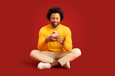 Foto de Sonriente hombre rizado afroamericano con teléfono inteligente en sus manos sentado en la pose de loto aislado en rojo. Chico carismático utilizando la aplicación móvil, mensajería de chat en línea, pasa tiempo en las redes sociales - Imagen libre de derechos