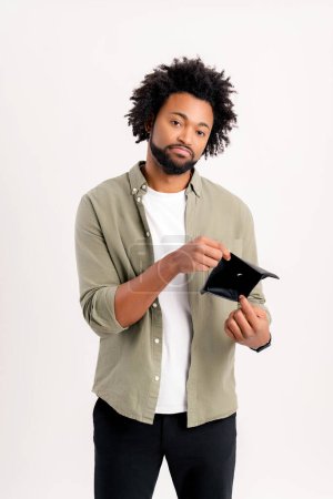 Photo for Depressed African-American young man is showing empty wallet isolated on white background, upset curly guy has lack of money, difficult financial situation due to the economic crisis - Royalty Free Image