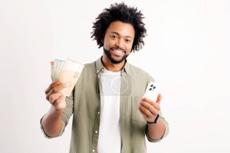 Téléchargez les photos : Earning online. Happy African-American man in casual wear holding cash money and smartphone standing isolated on white, guy won in online lottery, withdraw money from stocks, trader works on distance - en image libre de droit