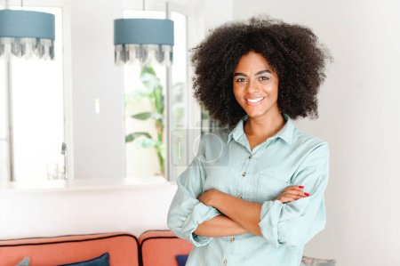 Téléchargez les photos : Femme ethnique regardant la caméra et souriant debout dans un appartement moderne, se sentant confort et détente. Joyeux afro-américain 20s jeune femme avec les bras croisés - en image libre de droit