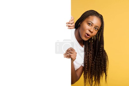 Téléchargez les photos : Funny astonished african woman peeps out from behind empty white banner, hiding behind huge paper poster, girl with pigtails surprised with deal, advertising, mock up, copy space, isolated on yellow - en image libre de droit