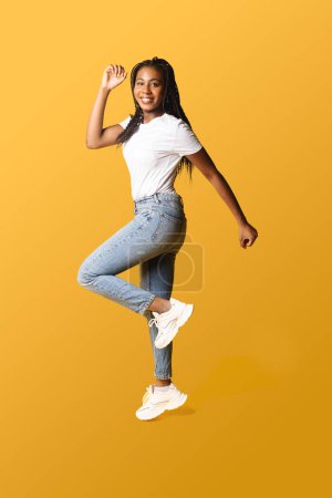 Téléchargez les photos : Full length photo of happy young 20s african woman with pigtails in white basic t-shirt and jeans jumping high, inspired by sale or best promotion, copy space, isolated on yellow - en image libre de droit