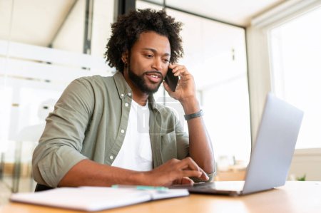 Téléchargez les photos : Jeune homme afro-américain utilisant un ordinateur portable sur le lieu de travail et a une conversation téléphonique, employé de bureau masculin bavarder sur le smartphone tout en tapant sur un ordinateur portable - en image libre de droit