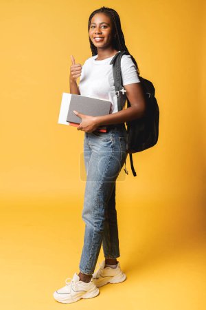 Foto de Foto de larga duración de una estudiante sonriente positiva con una mochila de pie aislada en amarillo, llevando libros y sembrando el pulgar hacia arriba, lista para estudiar, concepto de regreso a la escuela - Imagen libre de derechos