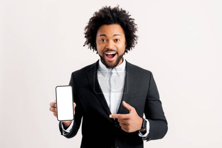 Foto de Incredible promotion. Surprised excited african-american 30s businessman wearing formal suit pointing finger at phone with empty screen isolated, male white collar worker presenting, advertising, ad - Imagen libre de derechos