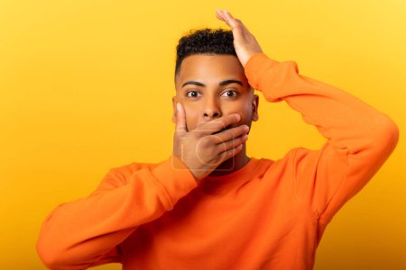 Photo for Excited shocked man closed mouth with hands, looking at camera, shocked bemused with news, win at lottery. Indoor studio shot isolated on yellow background - Royalty Free Image
