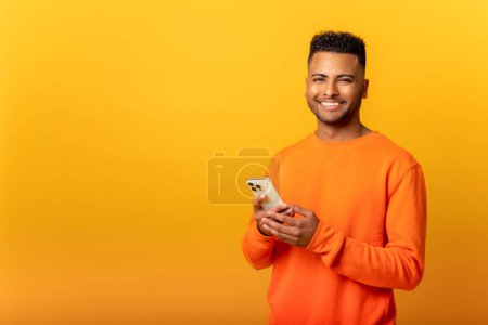 Photo for Photo of handsome cheerful smiling man holding mobile phone in the hand and smiling, isolated over yellow background, chatting using smartphone in front camera - Royalty Free Image