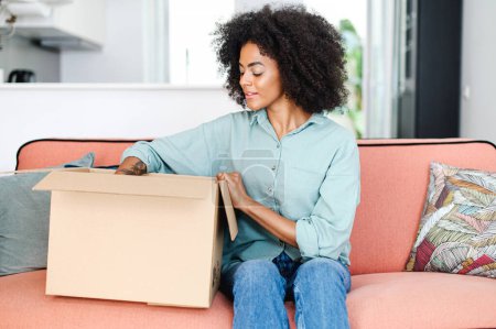 Foto de Mujer joven interesada con afro peinado desempacar paquete sentado en el sofá en el interior del apartamento, ama de casa alegre mirando dentro de la caja de cartón entregado a la puerta - Imagen libre de derechos