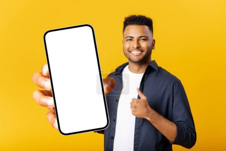 Foto de Cheerful Indian man with smartphone in the hand isolated on yellow background, showing smartphone with empty screen and thumbs up, advertising mobile app, showing deal - Imagen libre de derechos