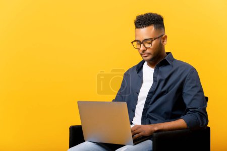Téléchargez les photos : Concentrated indian guy using laptop in casual shirt and glasses using laptop sitting isolated on yellow background, freelancer guy is working remotely, developer coding, student studying on distance - en image libre de droit