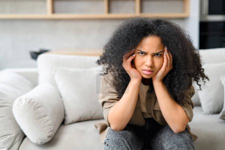 Téléchargez les photos : Femme multiraciale frustrée assise sur le canapé dans le chagrin, femme déçue se sent désespérée, inquiète, dépression, solitude, souffrant de forts maux de tête - en image libre de droit