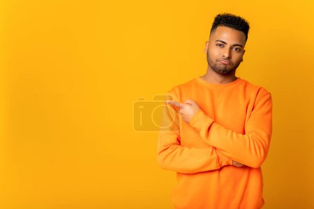 Foto de Sad young man points aside and looking at the camera, guy presenting novelty, advertising, isolated on yellow wall - Imagen libre de derechos
