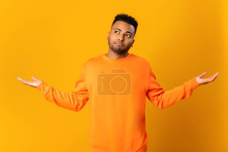 Photo for I dont know. Portrait of confused handsome young man standing with raised arms and looking at camera with answer. Indoor studio shot, isolated on orange background - Royalty Free Image