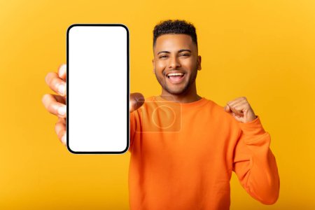 Photo for Young indian man demonstrates smartphone with blank screen and rejoicing, guy likes mobile app, looking and smiling at the camera, feels satisfied, isolated - Royalty Free Image