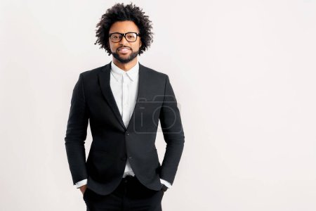 Photo for Smiling curly businessman standing with arms in pockets, looking at camera isolated on white. Young positive male entrepreneur in black formal suit. Proud successful multiracial small business owner - Royalty Free Image