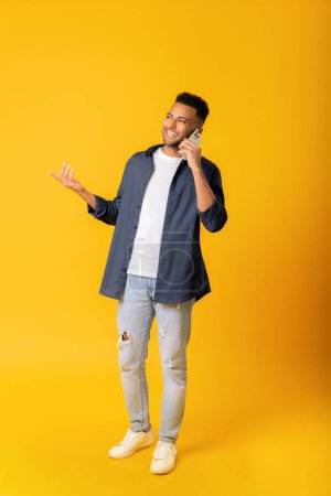 Photo for Overjoyed Indian guy talking on the smartphone over yellow background. Multiracial man enjoying phone conversation, glad to talk on the cell phone, received news. Copy space, advertising, full length - Royalty Free Image