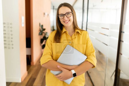 Photo for Smiling confident woman in eyeglasses looking at camera and standing in an office hallway. Portrait of confident businesswoman with a laptop, freelancer woman in coworking space - Royalty Free Image