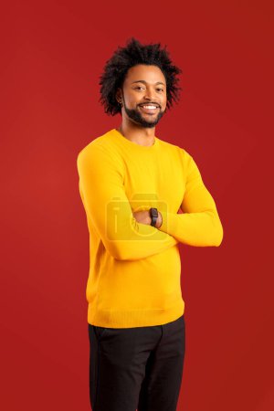 Téléchargez les photos : Souriant jeune homme afro-américain en vêtements décontractés debout avec les bras croisés confiant et heureux, regardant la caméra avec un large sourire dentaire isolé sur fond rouge - en image libre de droit