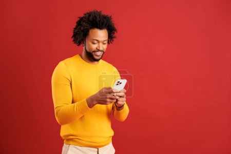 Téléchargez les photos : Portrait d'un homme barbu attrayant utilisant un téléphone portable avec une expression heureuse, accro au smartphone, textos sur les réseaux sociaux. Studio intérieur tourné isolé sur fond rouge - en image libre de droit