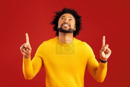 Foto de Hombre atractivo con expresión facial feliz apuntando hacia arriba con ambos dedos, presentando espacio de copia. Estudio interior plano aislado sobre fondo rojo - Imagen libre de derechos