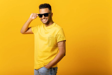 Photo for Overjoyed Indian man wearing sunglasses looking at the camera, smiling positively, being in high spirit. Carefree hispanic guy in yellow t-shirt isolated, having summer mood - Royalty Free Image