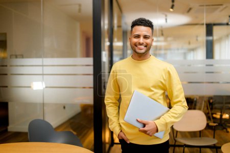 Photo for Smiling young indian businessman, arab male student, eastern employee, manager, coworker wearing casual yellow pullover standing carrying laptop, looking at the camera standing in office - Royalty Free Image