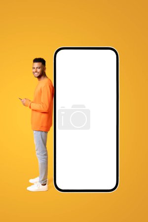 Foto de Sonriente chico atractivo de pie cerca de un enorme teléfono inteligente con la pantalla vacía en blanco, el hombre con el teléfono en sus manos la publicidad de la nueva aplicación o sitio web, maqueta - Imagen libre de derechos