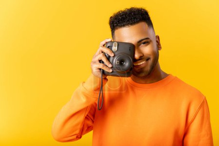 Photo for Smile. Cheerful glad Indian guy in casual t-shirt taking photo of you, holding instant photo camera and looking at the camera isolated on yellow, happy guy smiling with wide toothy smile. Studio shot - Royalty Free Image