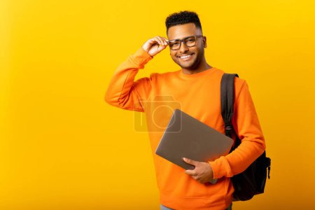 Téléchargez les photos : Intelligent intellectuel indien employé masculin ou pigiste homme tenant ordinateur portable et regardant la caméra avec sourire heureux, isolé sur fond jaune, étudiant arabe avec sac à dos ajustant ses lunettes - en image libre de droit