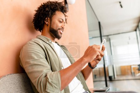 Téléchargez les photos : Portrait d'un employé masculin afro-américain concentré utilisant un smartphone, un homme d'affaires souriant répond aux e-mails, un homme indépendant travaillant sur la distance assis dans un espace de coworking, vue latérale - en image libre de droit