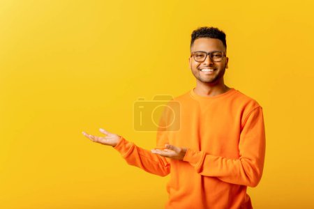 Photo for Cheerful indian man pointing at copy space with his palms, empty place for idea presentation, product advertising. Indoor studio shot isolated on yellow background - Royalty Free Image