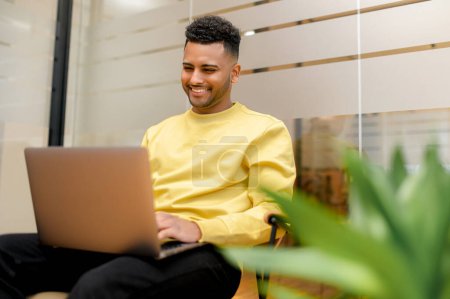 Foto de Alegre guapo empleado de oficina masculino usando el ordenador portátil en el espacio de coworking contemporáneo, estudiante árabe sonriente escribiendo correos electrónicos sentados en el banco en el pasillo, navegar por la web, preparándose para los exámenes - Imagen libre de derechos