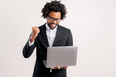 Téléchargez les photos : Homme d'affaires excité regardant l'écran de l'ordinateur portable et se réjouissant. Heureux afro-américain gars employé de bureau masculin dans la forme et les lunettes porter regarder fixement moniteur amusé avec une occasion inattendue - en image libre de droit
