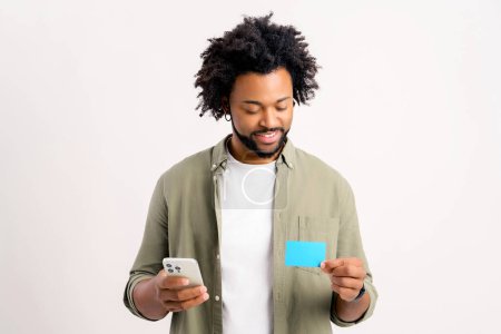 Téléchargez les photos : Béni jeune homme multiracial tenant la carte de débit et smartphone isolé. Homme chanceux en vêtements décontractés recevoir de l'argent des achats en ligne - en image libre de droit