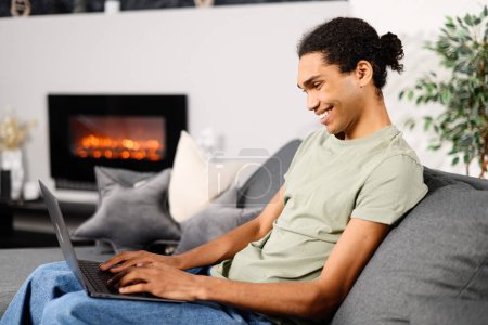 Téléchargez les photos : Vue latérale du jeune homme multiracial utilise un ordinateur portable pour un appel vidéo à la maison, il regarde la webcam avec plaisir sourire. Communication vidéo, zoom - en image libre de droit