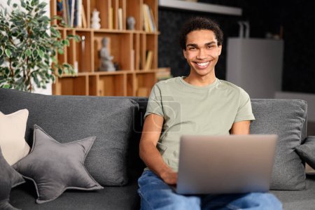 Photo for Handsome young multiracial man with beard sitting on the sofa with laptop at home, searching in the internet, watching film or serial, remote meeting, on the weekend, relaxed, listening performance - Royalty Free Image