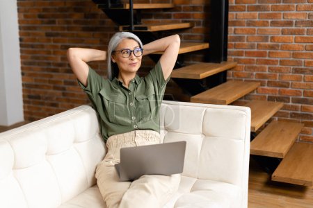 Photo for Modern mature 60s female enjoy leisure weekend at home, browsing wireless internet on computer. Happy elderly woman sit on couch in living room and looking away with smile - Royalty Free Image